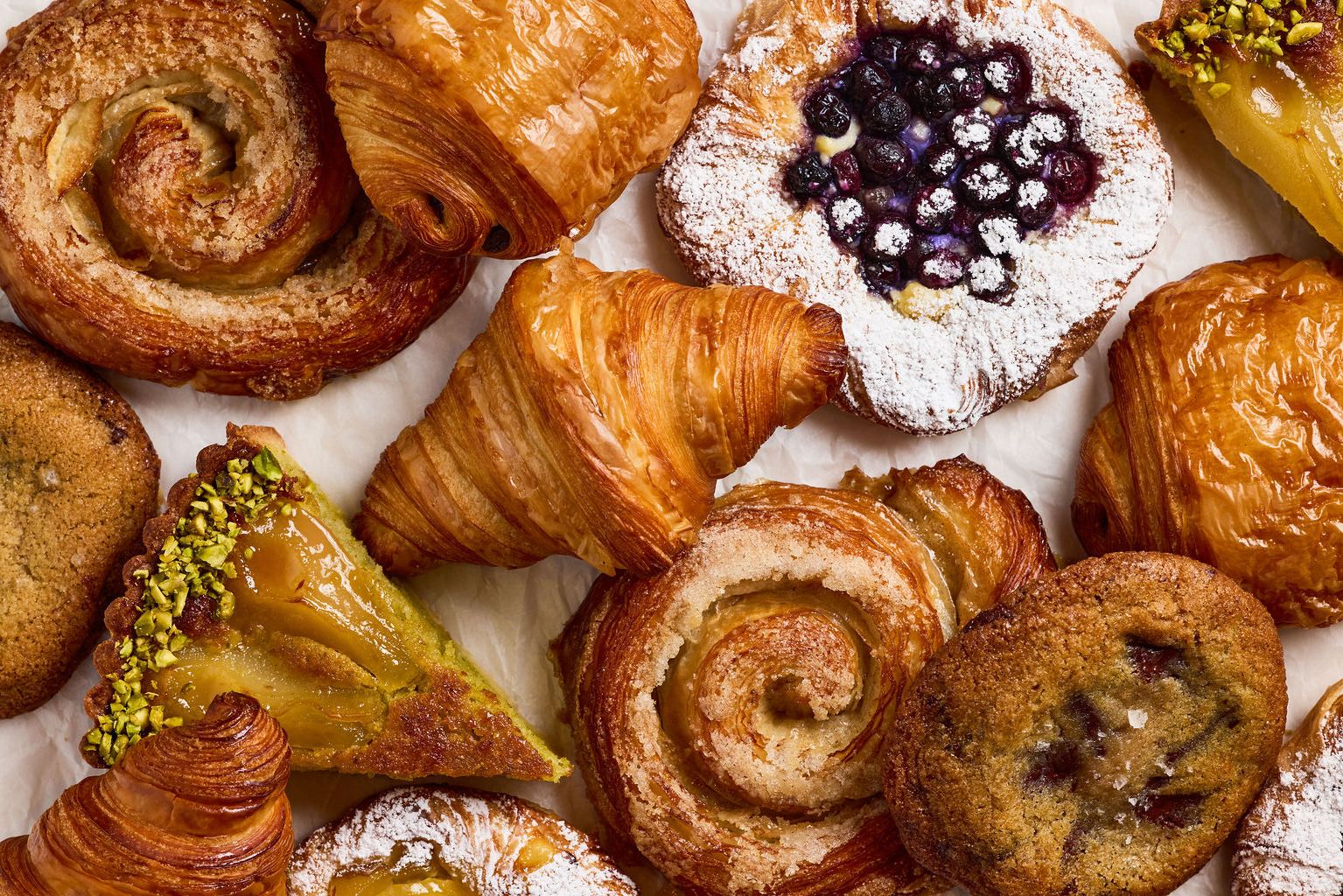 An overhead spread of Ciccio Bomba pastries — cinnamon rolls, croissants, blueberry tarts, pistachio frangipane.
